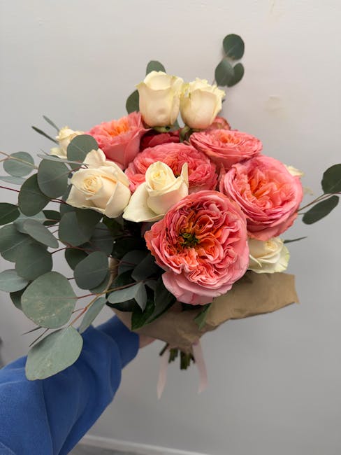 A bouquet of fresh floral arrangement featuring large, ruffled pink and peach ranunculus flowers, complemented by small white roses and sprigs of eucalyptus with rounded, matte green leaves. The flowe