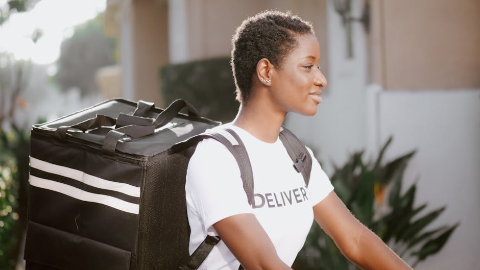 A female delivery person with short curly hair is walking outdoors, carrying a large black insulated delivery bag with white stripes on her back, in a residential area with blurred houses and greenery